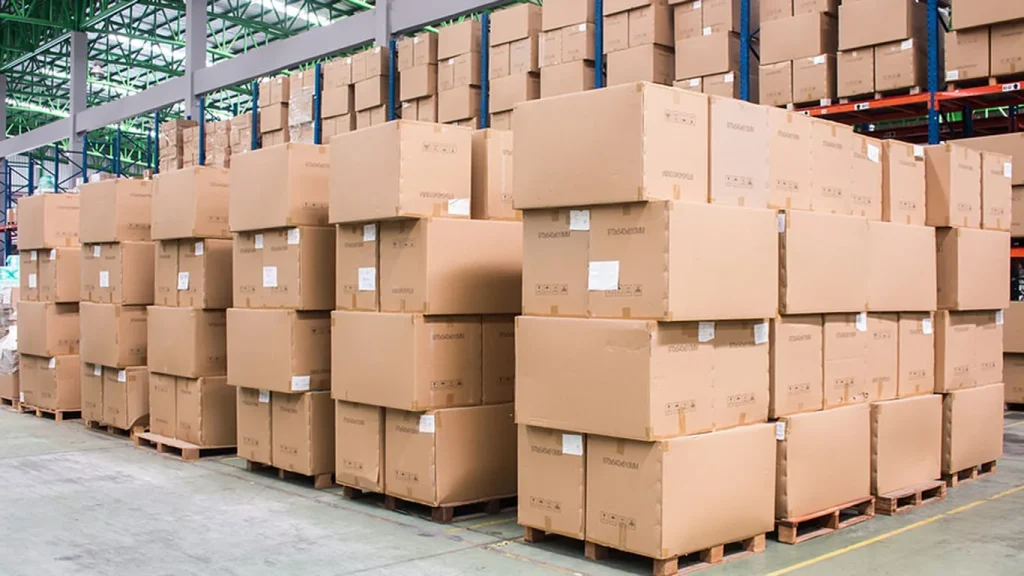 Rows of tall stacks of cardboard boxes on wooden pallets inside a large, modern Guangzhou 3PL warehouse with green and blue steel structural beams.