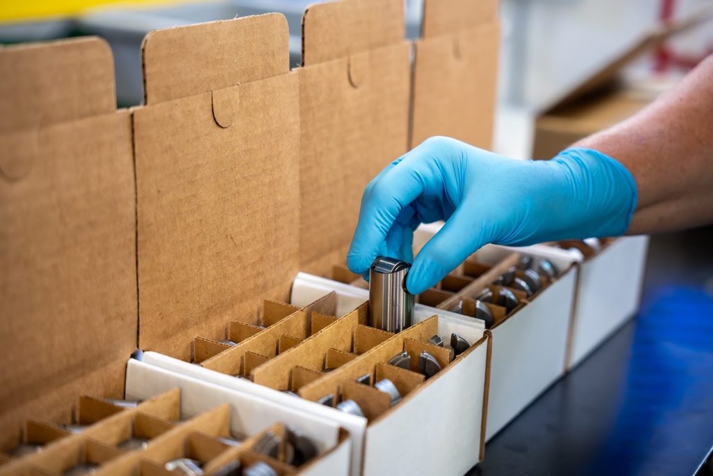 A worker wearing blue gloves carefully places a small metal component into a compartmentalized cardboard box for kitting and assembly.