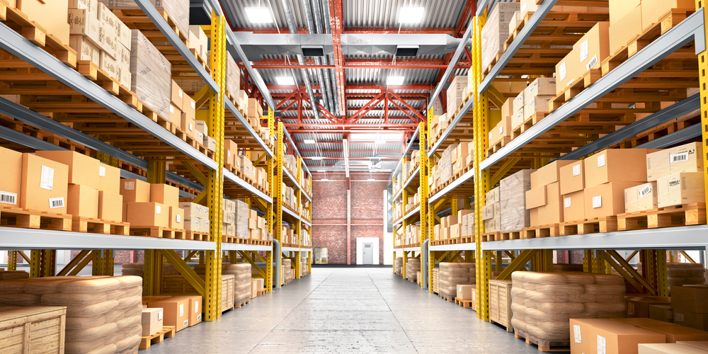 A wide view of a large warehouse aisle with tall yellow and blue pallet racks on both sides, fully stocked with cardboard boxes and goods on pallets.
