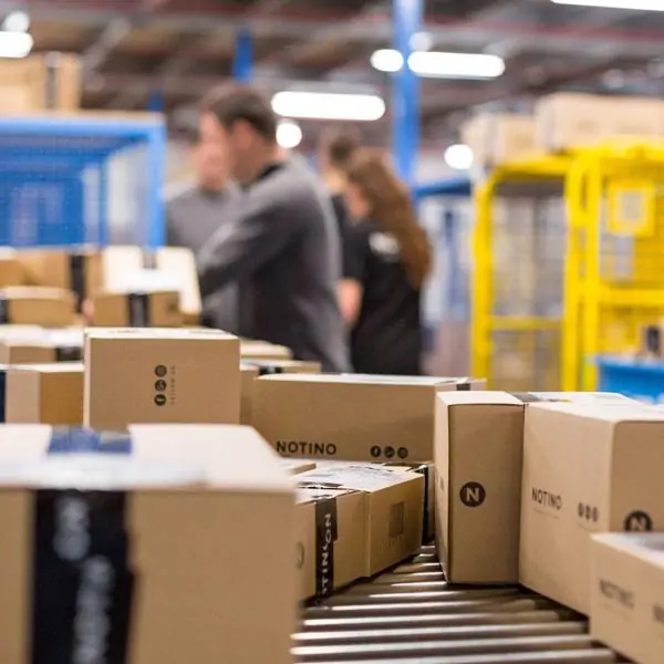 A warehouse conveyor belt carrying branded cardboard boxes labeled "NOTINO" while workers process orders in the blurred background.