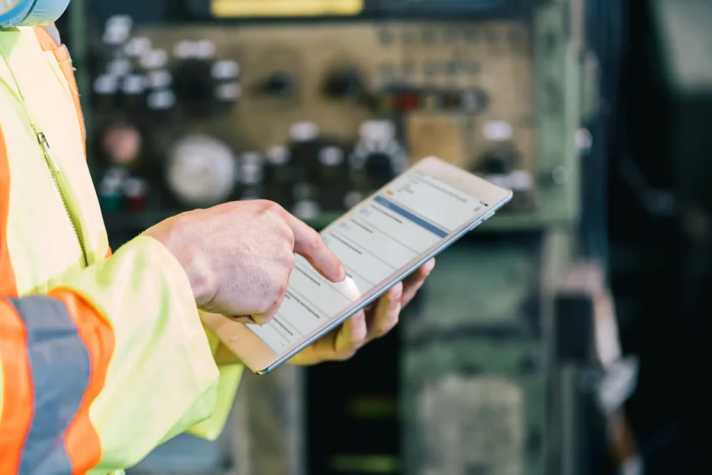 A close-up of a worker in a high-visibility vest using a tablet to perform a quality control (QC) check in an industrial setting.